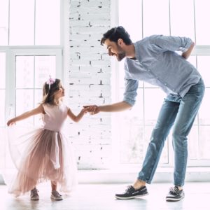A man and a young girl in a pink dress smile and hold hands while dancing together during the Daddy Daughter Recital Performance in a bright room with large windows.