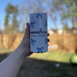 A hand holds up the 12 oz - Bow Tumbler, a white tumbler with blue bows and "Shining Stars Dance Company" text, set against a blurred outdoor backdrop with trees and a wooden fence.