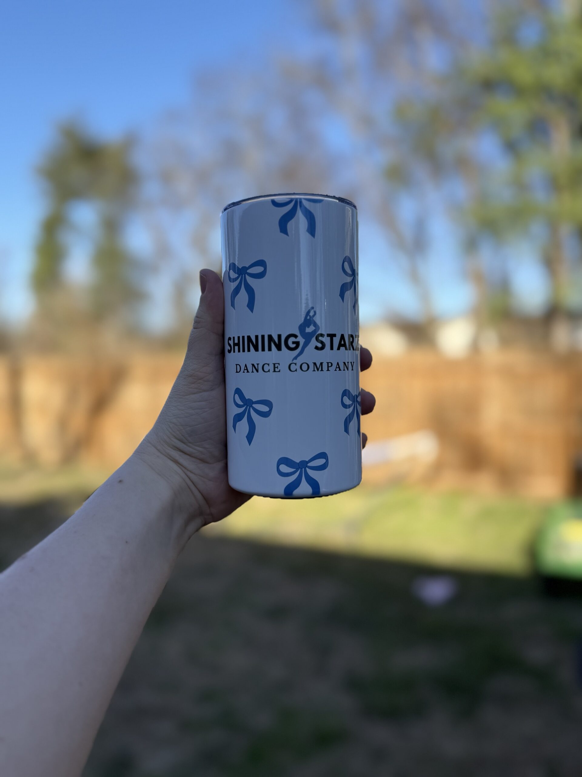 A hand holds up the 12 oz - Bow Tumbler, a white tumbler with blue bows and "Shining Stars Dance Company" text, set against a blurred outdoor backdrop with trees and a wooden fence.