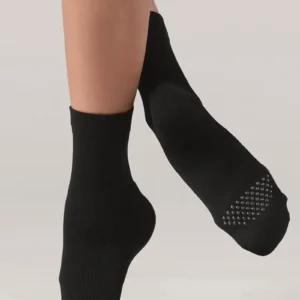 A person stands barefoot on a light background, displaying the bottom and side view of Weissman Grip Flow black crew socks with textured, non-slip grips on the sole.