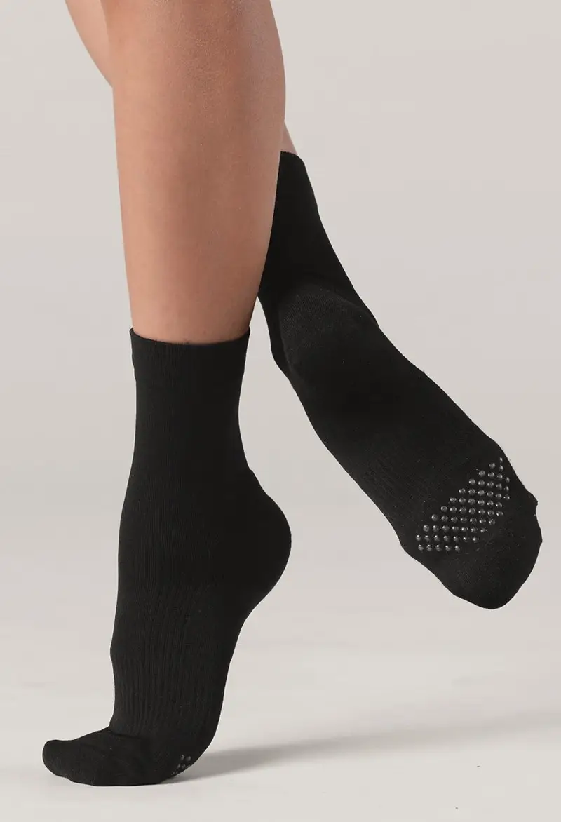 A person stands barefoot on a light background, displaying the bottom and side view of Weissman Grip Flow black crew socks with textured, non-slip grips on the sole.