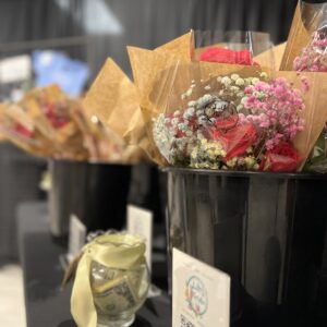 A close-up of colorful flower bouquets wrapped in brown paper, displayed in black buckets on a table. A small glass jar with dollar bills and a QR code sign are in the foreground. The background is softly blurred.
