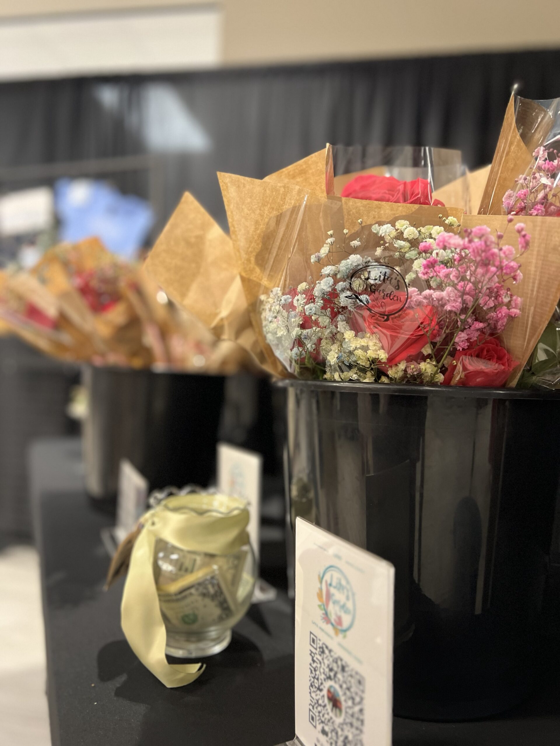 A close-up of colorful flower bouquets wrapped in brown paper, displayed in black buckets on a table. A small glass jar with dollar bills and a QR code sign are in the foreground. The background is softly blurred.